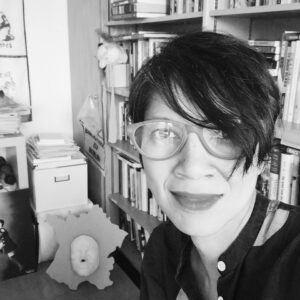 Black and white photograph of Roxanne Panchasi from the shoulders up, with a book shelf in the background