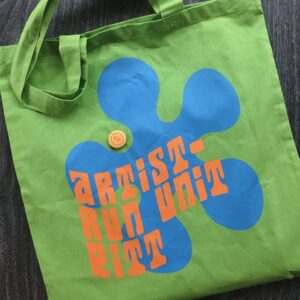 Photograph of a lime green tote bag against a dark background. Design of a blue bubbly flower motif with the orange text, "artist-run unit pitt". Small yellow and pink button with the UNIT/PITT logo.
