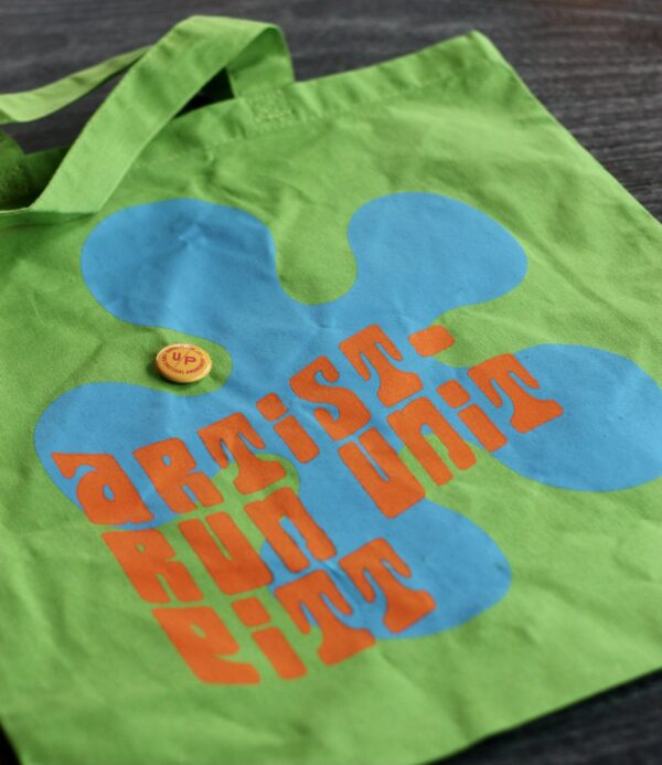 Close-up photograph of a Mystery Tote against a dark background. Green bag with blue flower motif and orange letters that read, "artist-run unit pitt," and a yellow and pink button with UNIT/PITT's logo