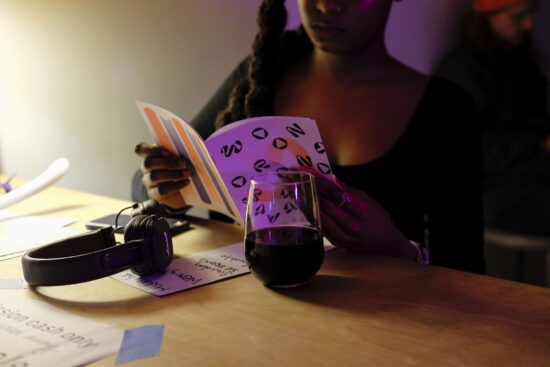Close-up photograph of a figure holding a colourful zine with the words "Wrong Wave" on the cover. Also in the photograph: a desk, headphones, a glass of what looks like wine. Low light. Photograph by Sara Baar