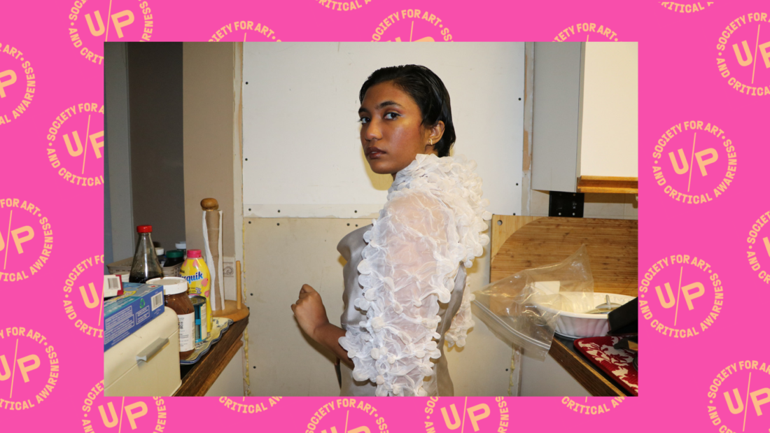 An image of Nadia wearing a puffy white dress with her shoulder to the camera. She is looking back at the camera. The photo is overlaid against a pink background overlaid with a pattern of peach UNIT/PITT logos arranged diagonally.An image of Nadia wearing a puffy white dress with her shoulder to the camera. She is looking back at the camera. The photo is overlaid against a pink background overlaid with a pattern of peach UNIT/PITT logos arranged diagonally.