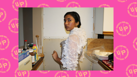 An image of Nadia wearing a puffy white dress with her shoulder to the camera. She is looking back at the camera. The photo is overlaid against a pink background overlaid with a pattern of peach UNIT/PITT logos arranged diagonally.An image of Nadia wearing a puffy white dress with her shoulder to the camera. She is looking back at the camera. The photo is overlaid against a pink background overlaid with a pattern of peach UNIT/PITT logos arranged diagonally.