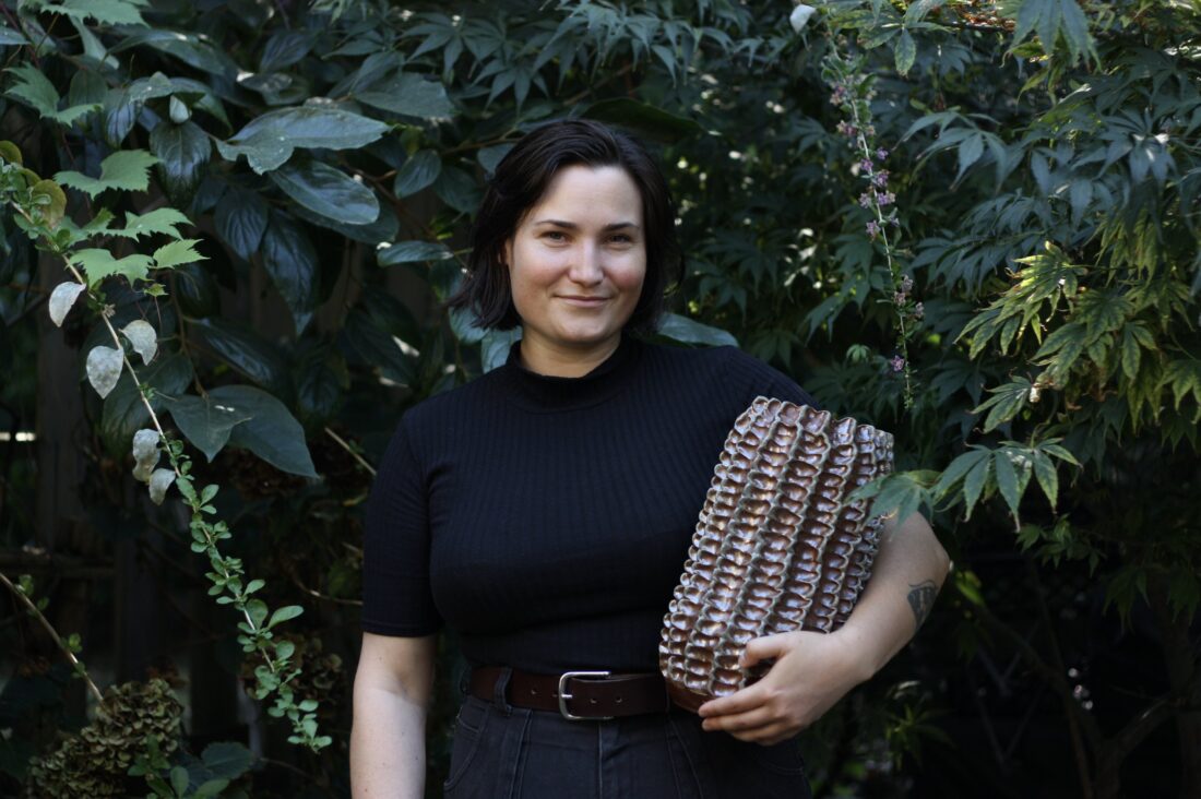 Photograph of a woman with short dark hair, light skin, smiling at the camera. She wears a black mock-turtle neck tee and is holding a large ceramic vessel that is copper-coloured with textures. The background is dark green foliage. Photograph of Brit Bachmann, 2022