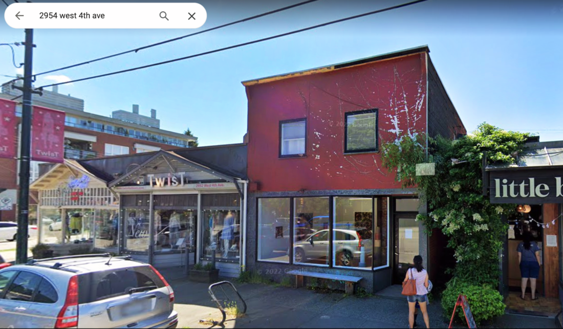 a google streetview image of 2954 west 4th avenue building in vancouver. the building is a burgendy-maroon colour with street level windows, and two smaller 2nd-floor windows. A woman is standing in front of the building, and there is a bike rack in front, as well as a bench for sitting on. singles torey building are on either side. a blue sky is in the background.