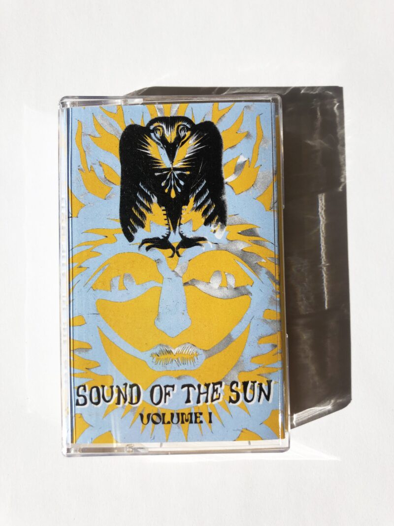 a light blue and yellow cassette tape on a white background. There is a black phoenix image overlaid on top of a cut-out "sun" face. text on cassette tape reads" SOUND OF THE SUN, VOLUME I