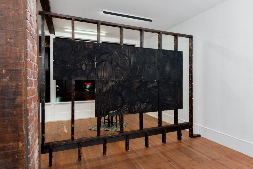 Installation image of Kiyoshi Whitley's work 'days; featuring a blackened piece of plywood, with carved linear forms, chalk, linseed oil, and designs burned onto the surface with a propane torch.