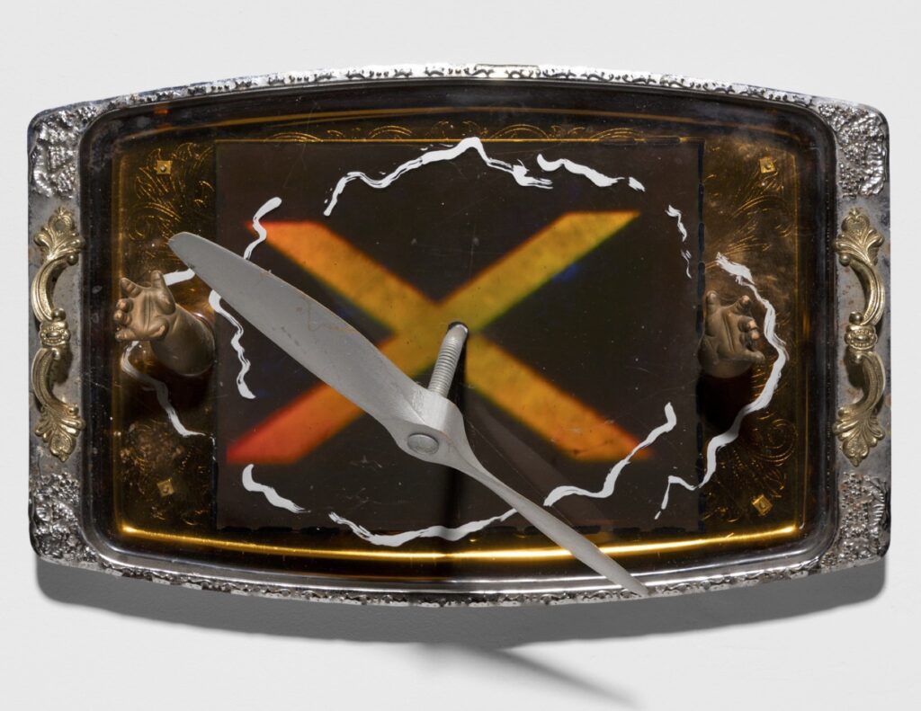 a holographic sculptural assemblage featuring a silver propeller emerging from a silver serving tray. bronze coloured baby hands emerge from the tray, with an orangey-red-and green X-shaped hologram in the center.