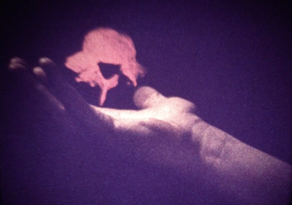 A dark purple, grainy film still with an outstretched hand appearing to hold a pink puff of smoke (it is a hologram)