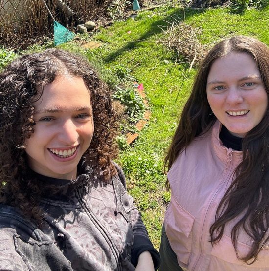 New Executive Director Alison Bosley (left) and former Executive Director Catherine de Montreuil (right) smiling in U/P's backyard garden. Ali is a woman with dark brown curly hair and blue eyes, wearing a bleach dyed sweater. Catherine is a woman with long straight brown hair and green eyes wearing a light pink zip-up windbreaker. They are standing close and smiling into the camera.
